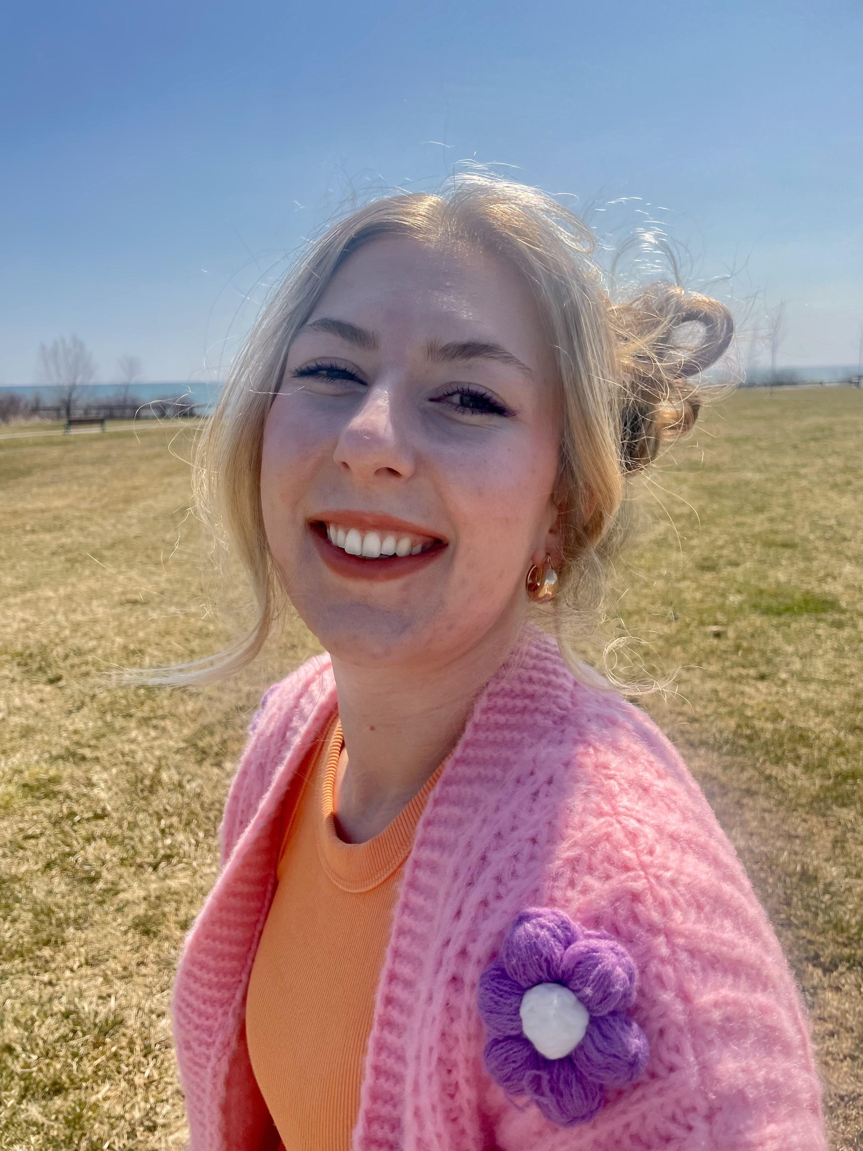 Twenty-something blonde woman smiling in a field. She's wearing an orange shirt and pink cardigan with crocheted flowers on it.